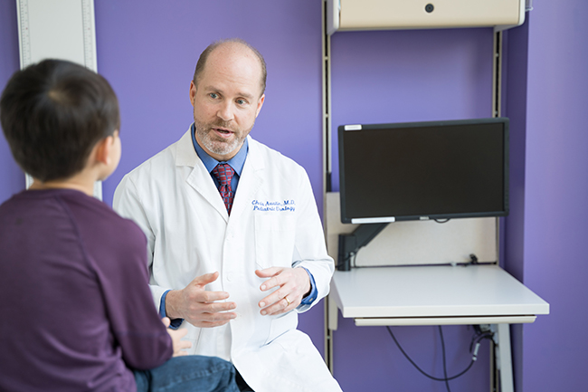 A male doctor sitting in an exam room providing an explanation to a little boy (a patient) who is sitting on an exam table.
