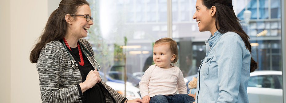 A female doctor and another woman standing on either side of a baby girl sitting on an exam table, all three smiling.