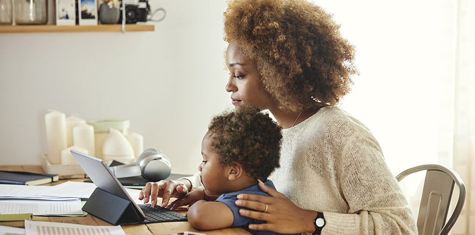 Photo of a person seated at a table reading a laptop while a child sits on their lap