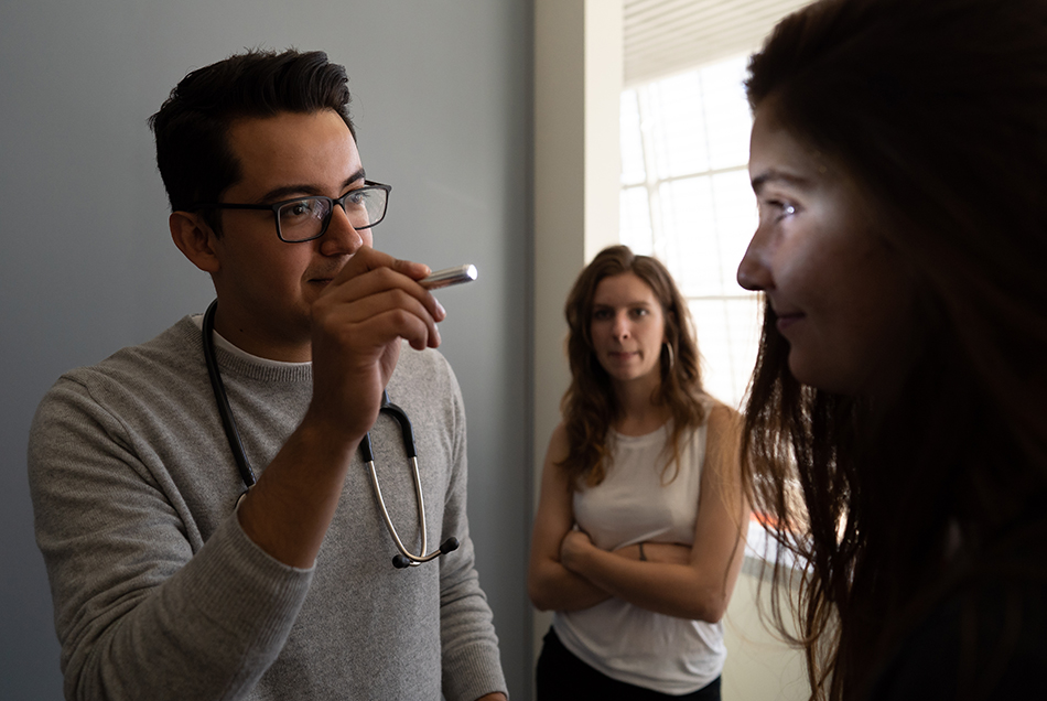 a medical student wearing glasses on his face and a stethascope around his neck using a pen light to examine the eyes of another medical student