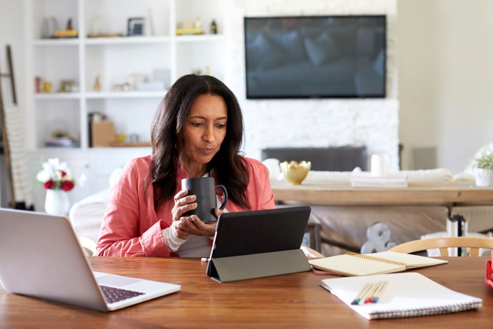 woman working at desk, with warm drink, in front of two computers