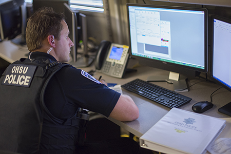 An OHSU police officer sitting at a desk staring at his computer screen and writing something down.