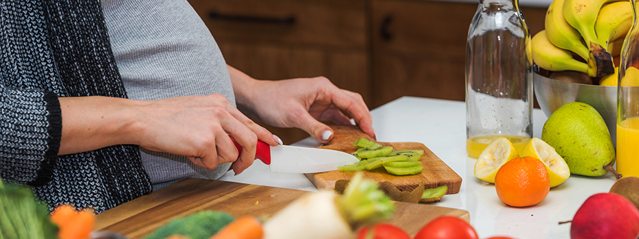A pregnant person cutting up a colorful variety of fruits and vegetables.