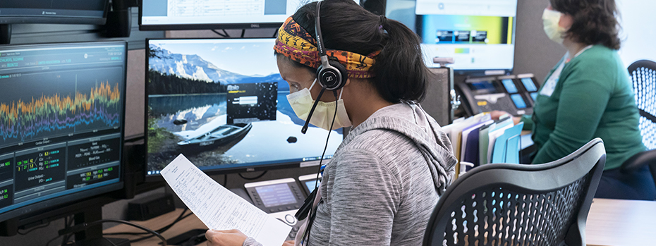 A virtual ICU nurse wearing a headset reviews a document and real-time patient data on her computer screen.