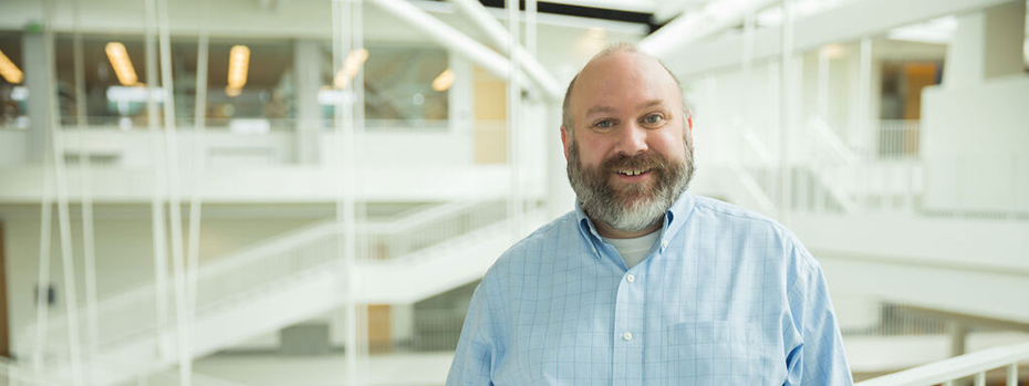A physician assistant stands in a big, open, interior space. He sports a beard and a grin. The mood is optimistic.