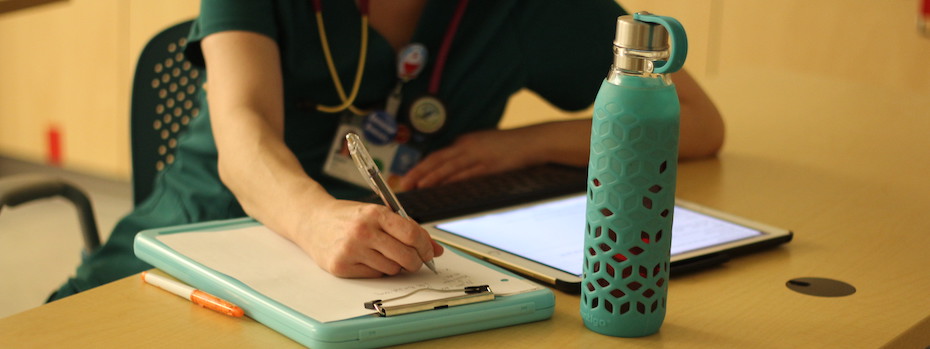 Close up of a student writing on a clipboard with a tablet and water bottle on the table.