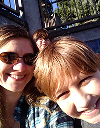 A woman smiling outside on a sunny day with a kid next to her and another behind her.