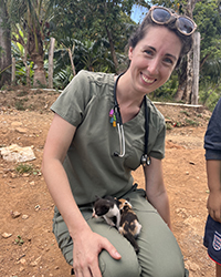 A woman crouching on the ground outside with a baby animal in her lap.