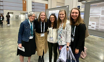 Five women smiling as they stand in front of a gallery of research posters at a conference.