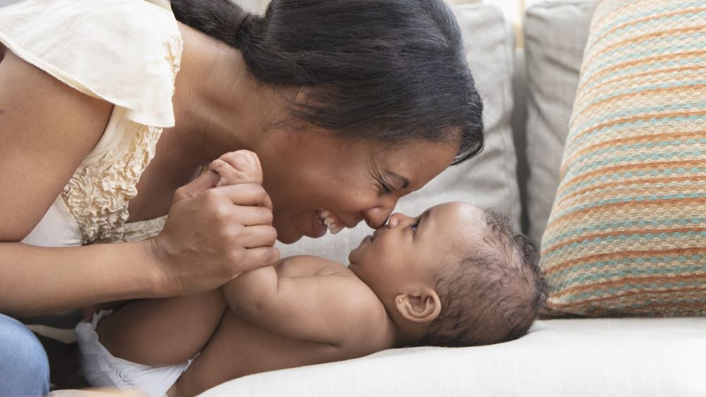 A mother smiles with her baby as they look at one another on a white couch.