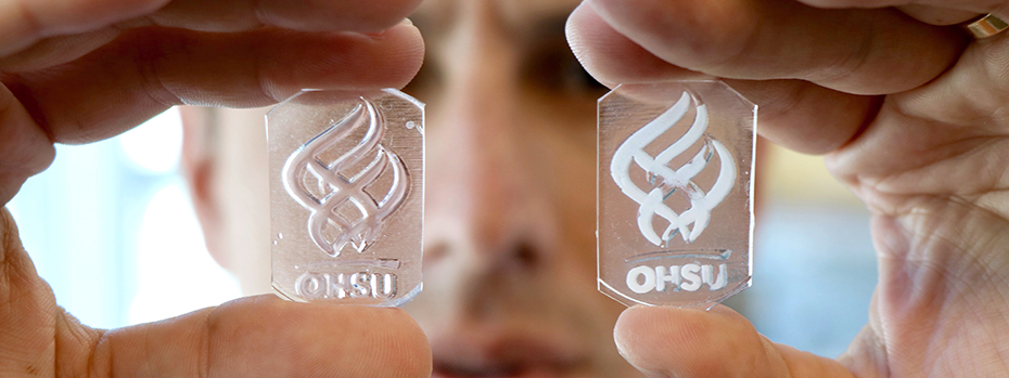 Hands holding up two small glass plates with opaque, engineered material in the OHSU torch logo. The researcher's face is in the background.