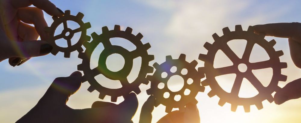 Two pairs of hands holding up four gears against a sunny sky.