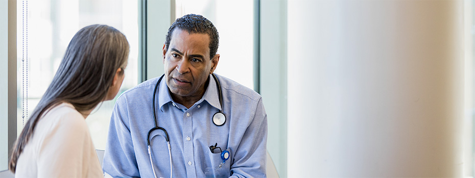 A student wearing a button up shirt and a stethoscope around their neck speaks with a patient.