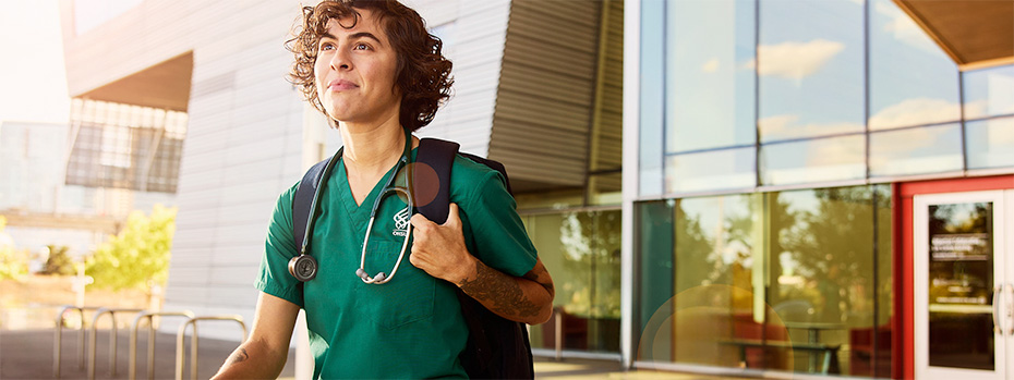 An OHSU nursing student in green scrubs with a stethoscope and backpack walks outside the Robertson Life Sciences Building in Portland, Oregon.