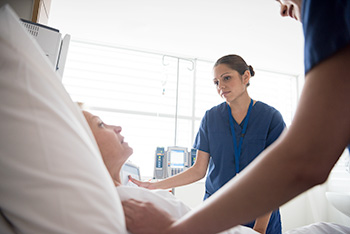An OHSU nursing student in blue scrubs stands at a patient’s bedside, listening attentively.