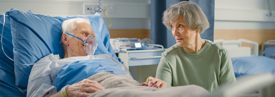 An elderly male patient wearing an oxygen mask rests in a hospital bed. He holds hands with and smiles at a woman visiting him.