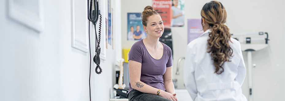 A young female patient sits on an exam table talking to a female doctor.