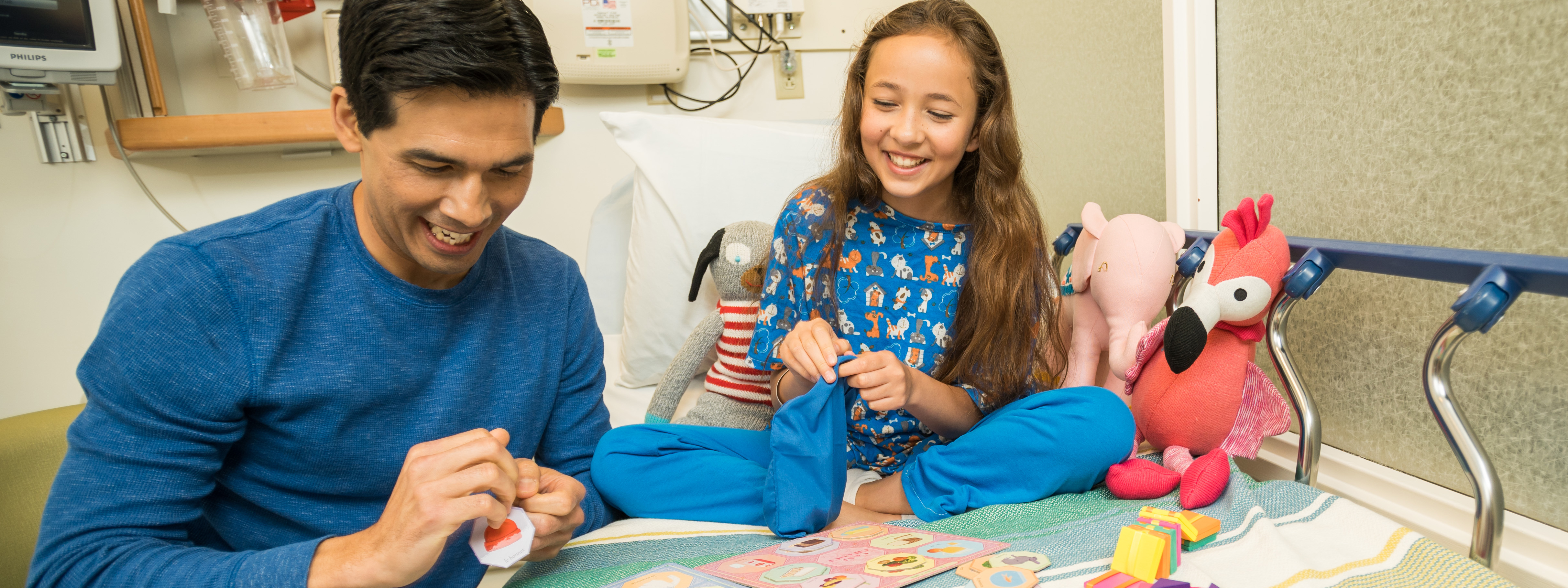 A young girl sits on a hospital bed in Doernbecher Children's Hospital, playing games with her dad who is visiting her.