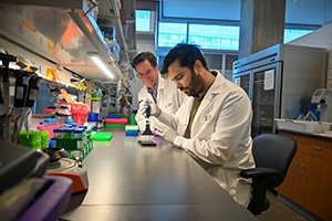 Two men wearing OHSU-branded lab coats sit in a research lab, with one handling a specimen as the other watches.