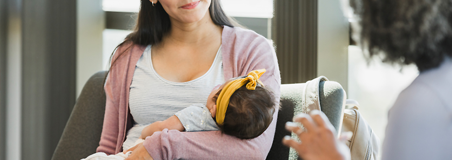 Post-partum woman holds her baby while talking with a care provider in a clinic. The photo is cropped to center the baby so the patient and care provider are unrecognizable.