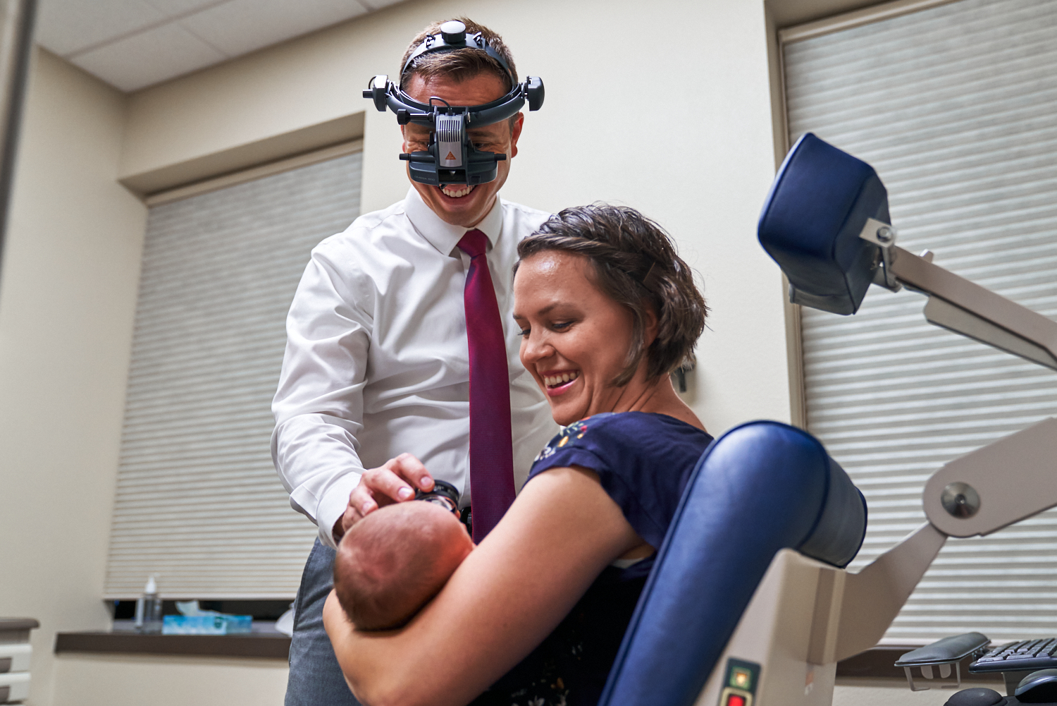 A doctor wearing a device over his face leans over a mother holder her baby so he can perform an eye exam.
