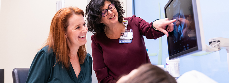 A female patient lying down on an exam table as two female providers stand beside her looking at an ultrasound monitor above the patient.