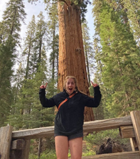 A woman in a forest standing in front of a tall tree pointing up at it.