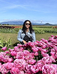 A woman sitting down smiling behind a patch of pink flowers with mountains visible behind her on a sunny day.