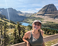 A woman leaning against a wooden railing with a river gorge and mountains visible behind her on a sunny day.
