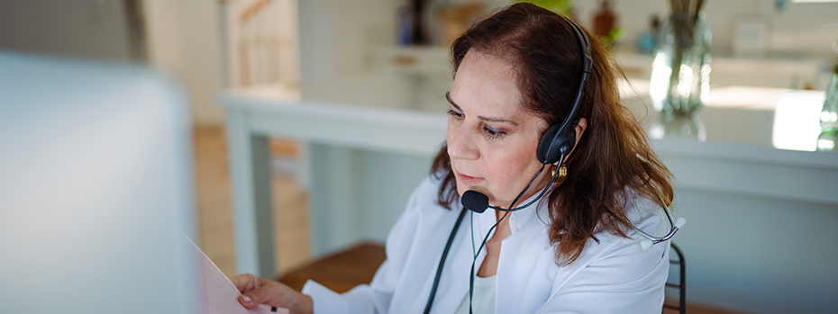 A woman sitting at a desk in front of a computer with a telephone headset on reviewing a patient chart.