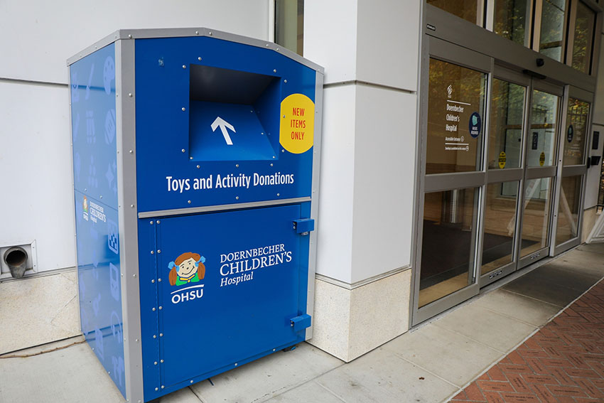 A large blue bin with a slot for placing toys and activity donations located outside the entrance to Doernbecher Children's Hospital. 