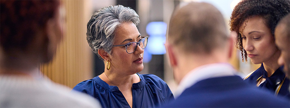  Educators engaged in collaborative discussion. A woman in glasses and navy blue attire speaks with colleagues in a professional setting.
