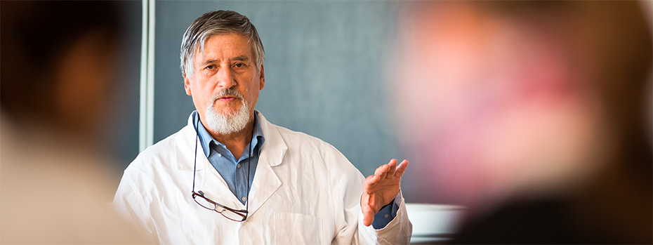 A medical educator in a white coat gestures while teaching, with students visible in the foreground out of focus.