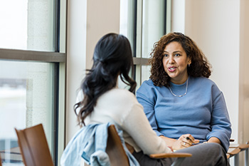 Two women sit across from one another while engaged in conversation in an office setting.
