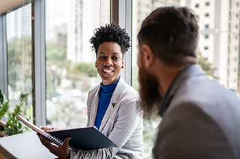 Two faculty members have a one-on-one meeting in an office setting. A woman holds a notebook while engaged in conversation with a colleague.