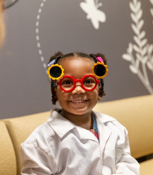 A preschool girl wears red and yellow sunflower glasses and a white doctor coat. She smiles at the camera.